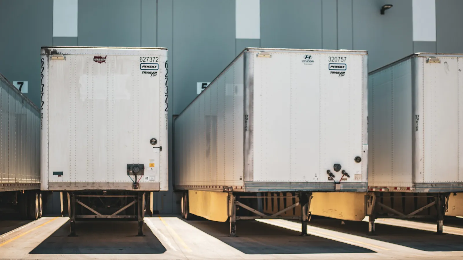 Three white semi-truck trailers parked at loading docks in a warehouse facility casting shadows on concrete.