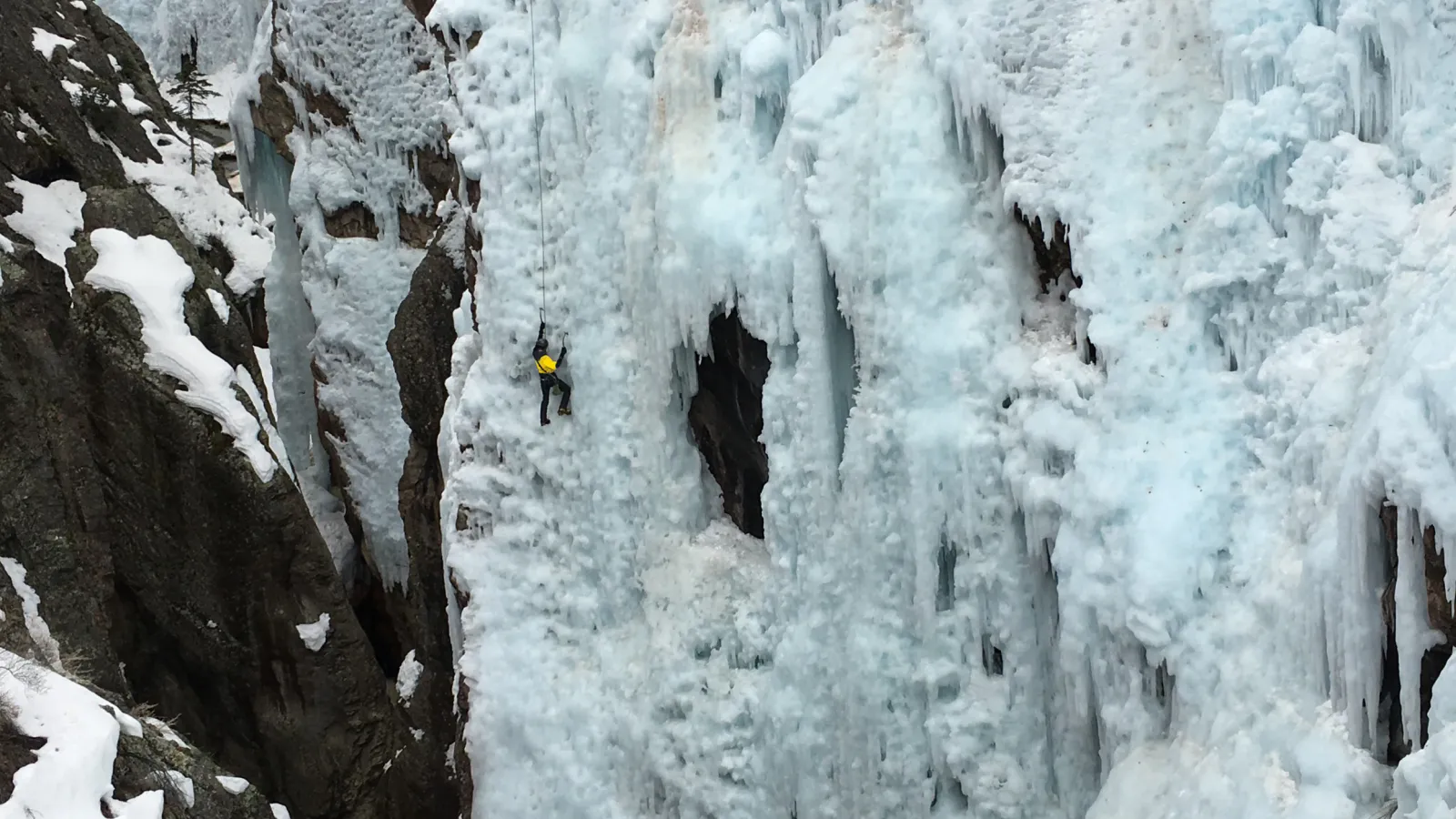 Climber in yellow jacket scaling a steep, frozen ice waterfall surrounded by snow-covered rocks.