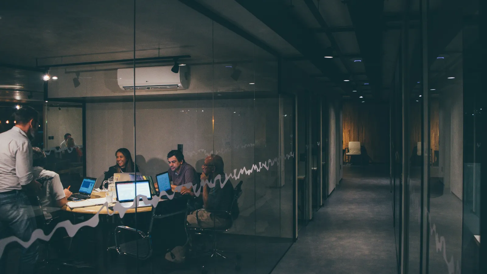Team of professionals collaborating in a modern glass-walled conference room with laptops and notebooks during evening.