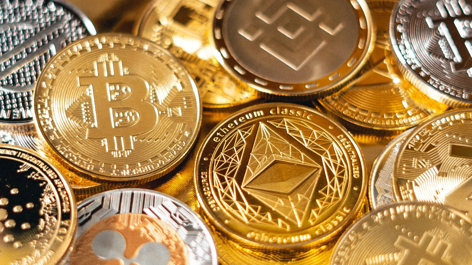 Close-up of assorted physical cryptocurrency coins including Bitcoin, Ethereum Classic, Binance Coin, and Ripple on gold surface.