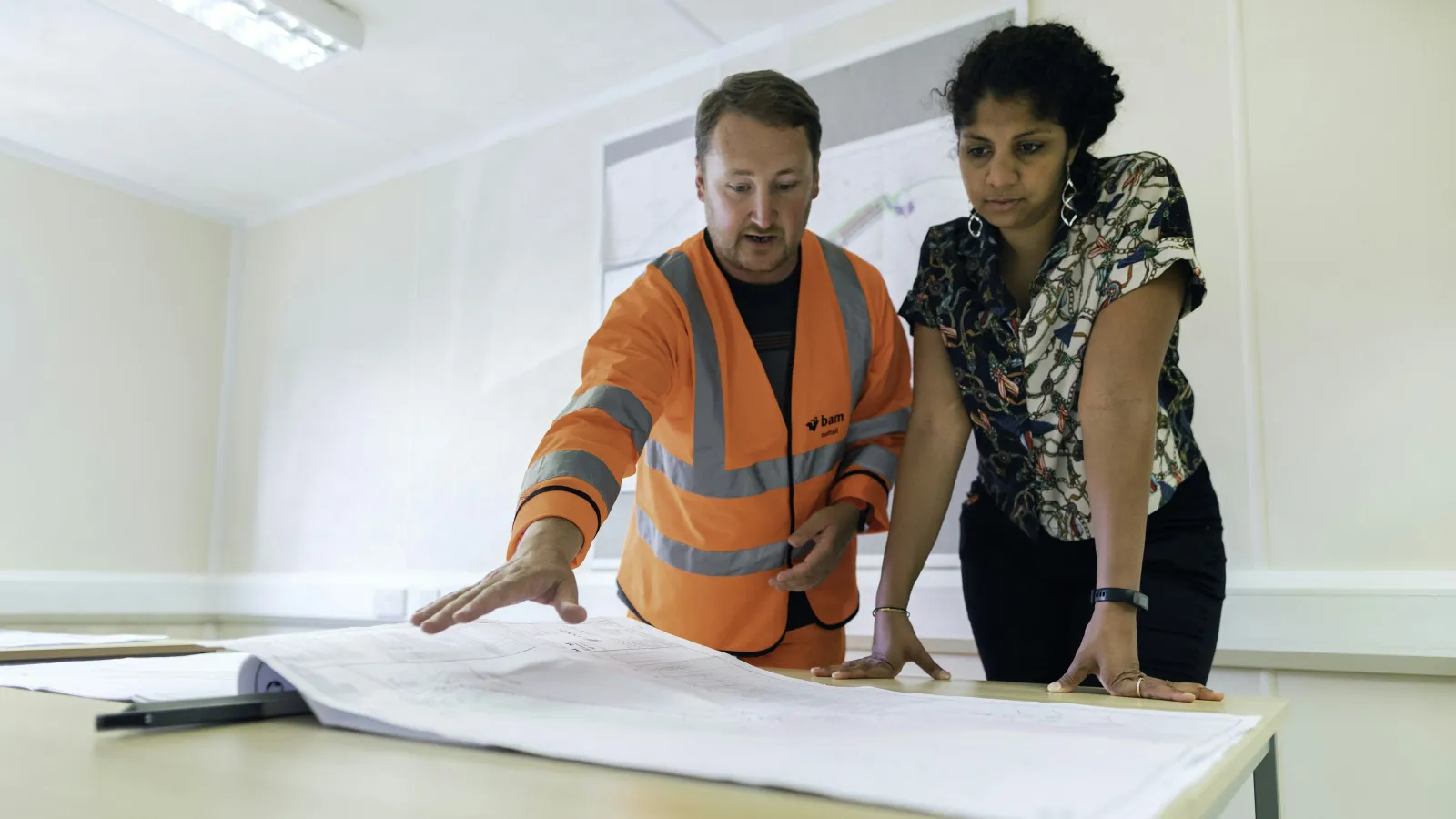 Man in orange safety vest and woman reviewing large architectural plans on table indoors