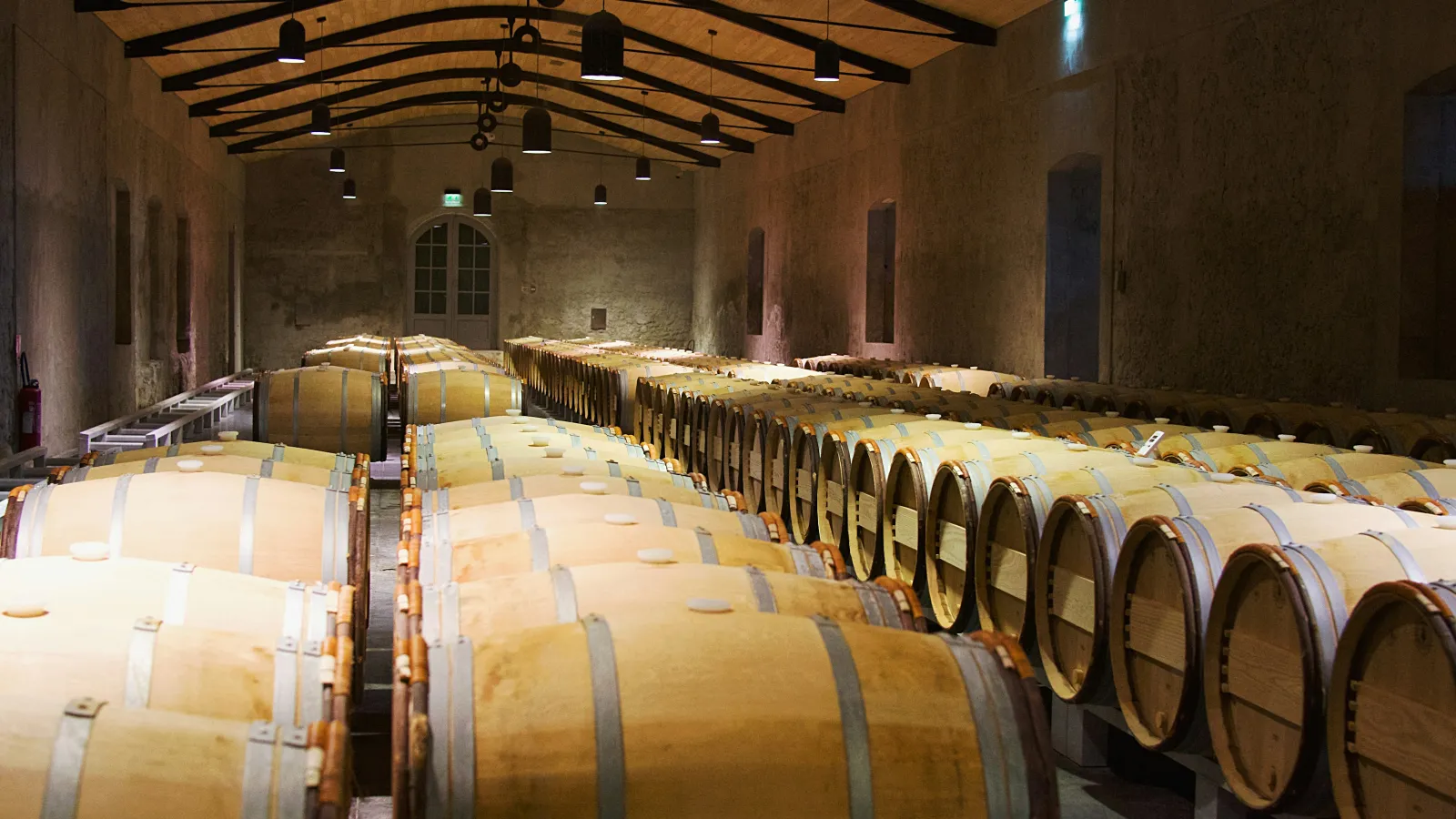 Dimly lit cellar with rows of wooden wine barrels aging in a rustic, arched stone room with warm lighting.