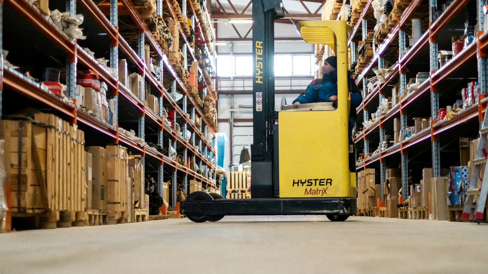 Yellow forklift operating between tall shelves stocked with wooden crates and boxes in a warehouse aisle.