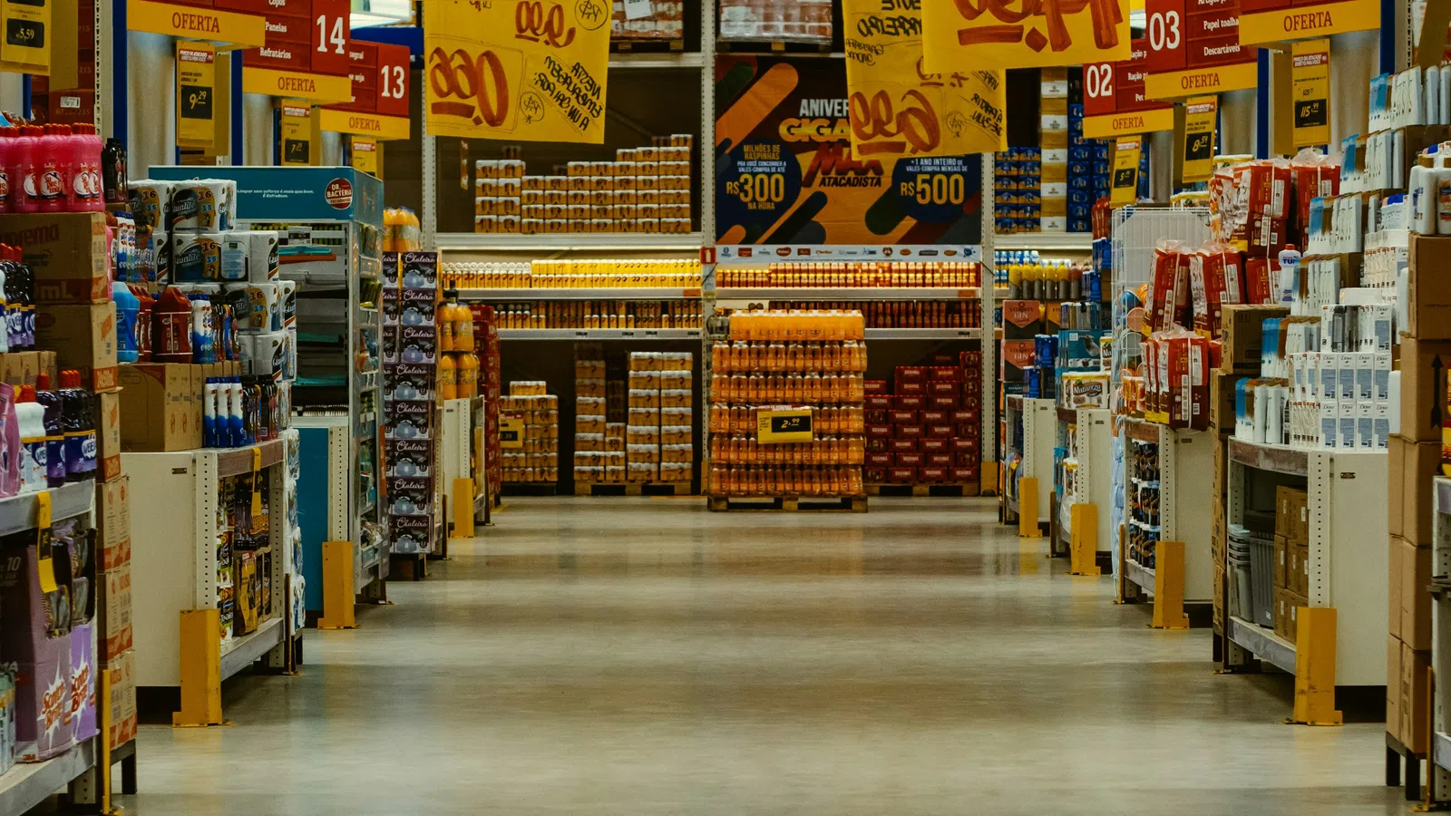 Wide aisle in a warehouse-style store with yellow price signs hanging from the ceiling and shelves stocked with products.