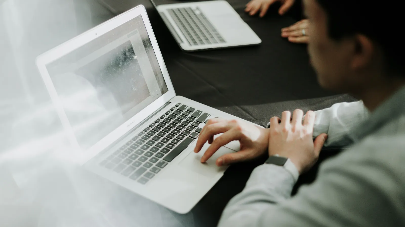 Person using a laptop at a black table with another laptop and hands visible in the background