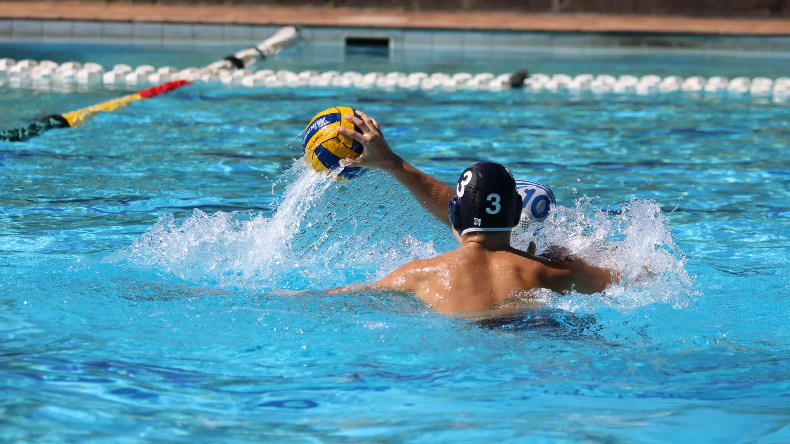 Water polo player wearing cap number 3 throws ball in outdoor swimming pool during match play.