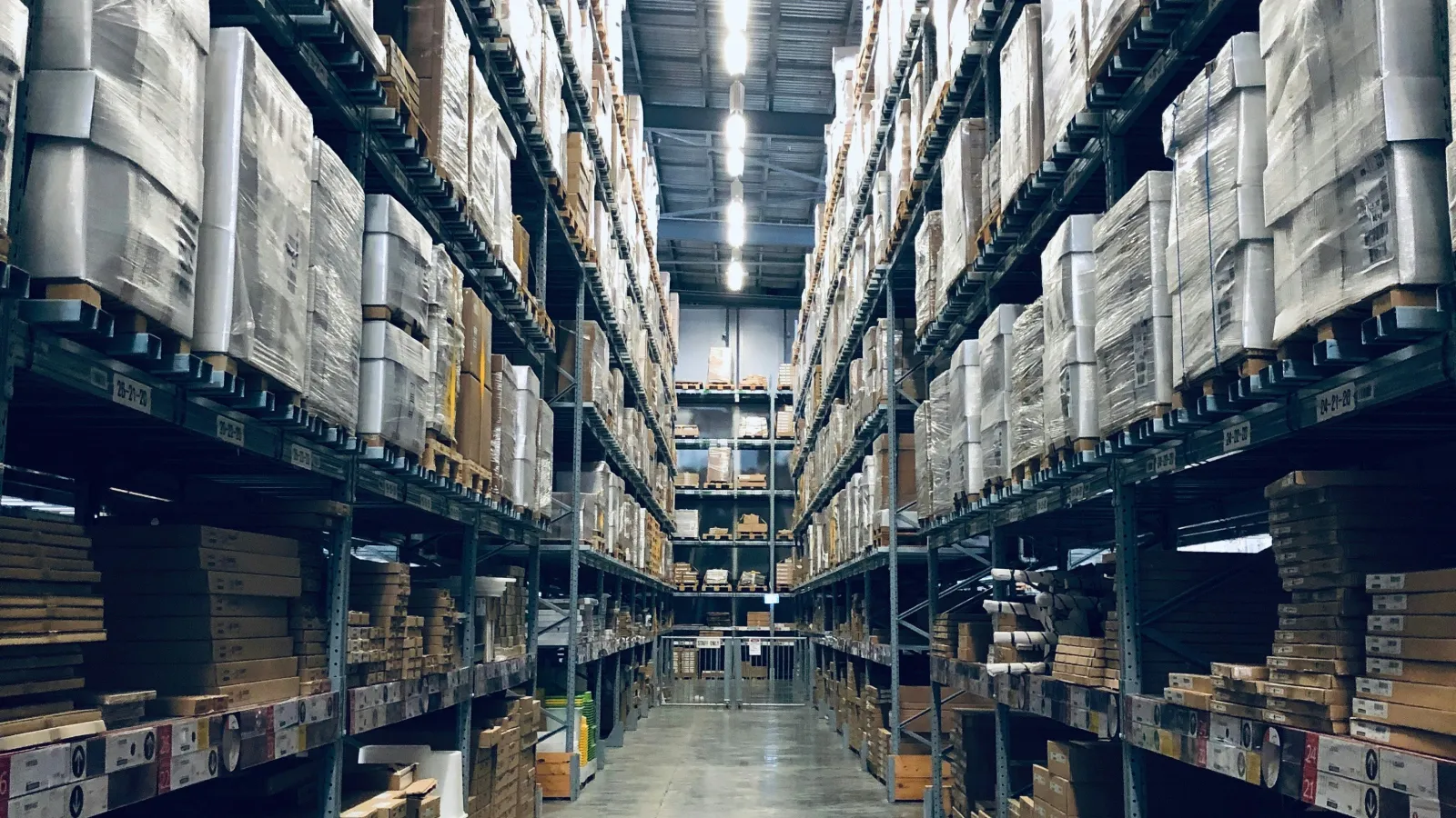 Long aisle in a large warehouse with tall shelves stacked with boxed goods and pallets wrapped in plastic.