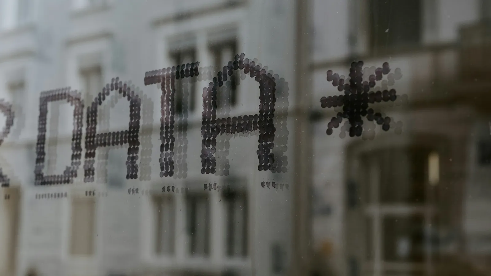 Close-up of a glass window with the word DATA* made of black dots and blurred buildings reflected behind it.