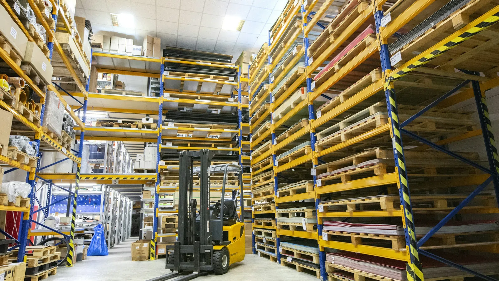 Warehouse interior with tall metal racks filled with pallets and a yellow forklift in the center aisle.