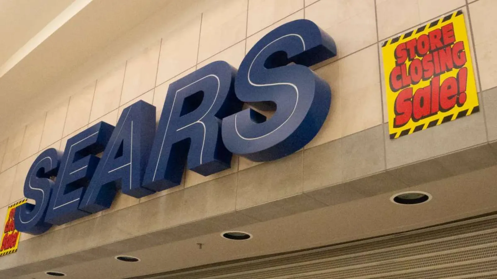 Sears store sign with bright yellow and red 'Store Closing Sale' signs on a beige wall.