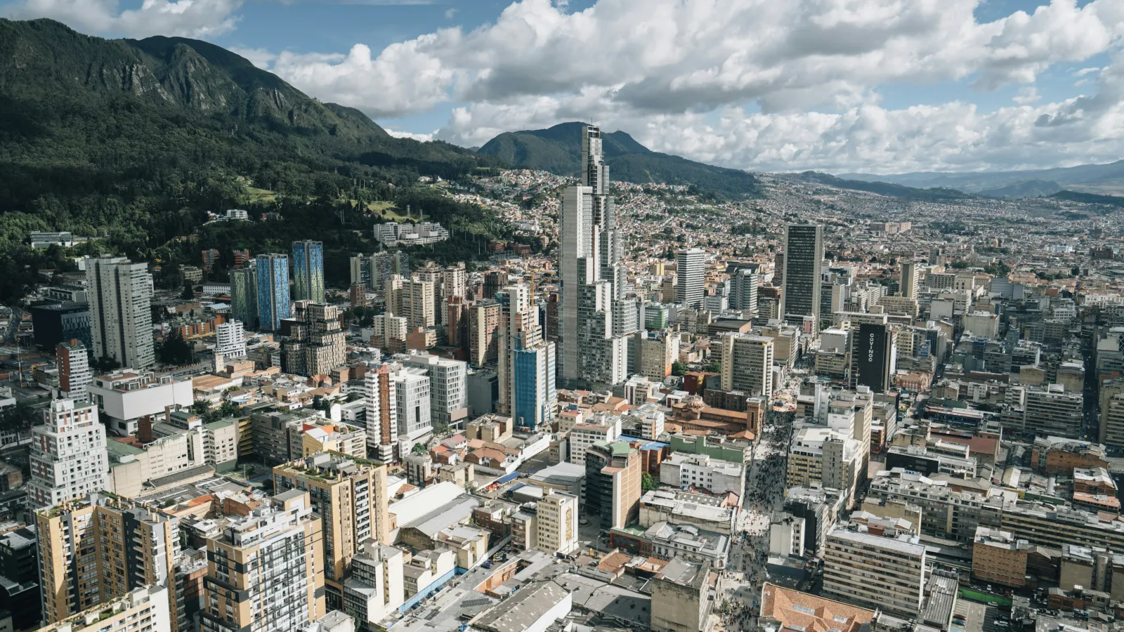 Aerial view of a city with tall skyscrapers surrounded by green mountains under a partly cloudy sky.