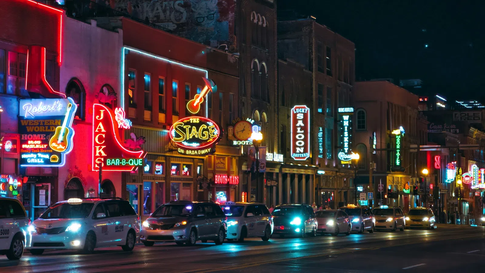 Nighttime city street filled with colorful neon signs and lined with parked cars.