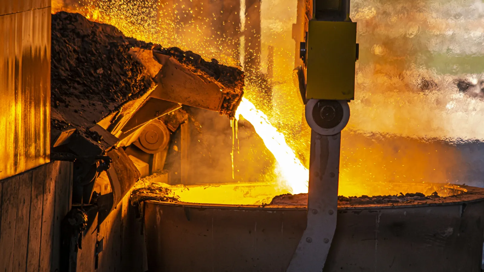 Molten metal pouring from a ladle into a large container in a bright industrial foundry setting.