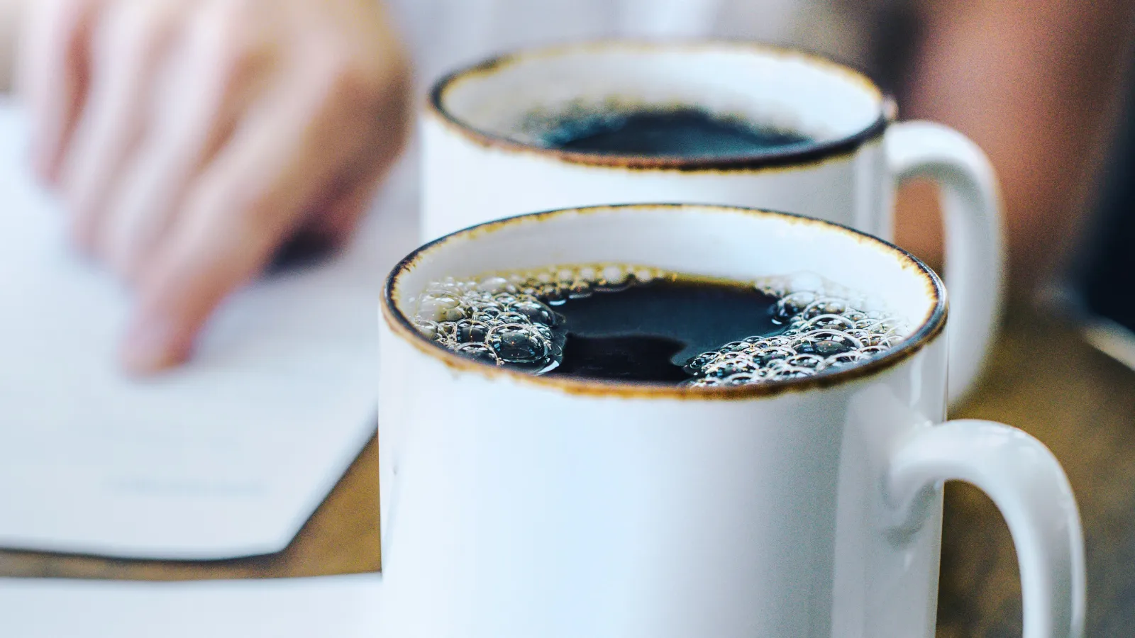Two white ceramic mugs filled with black coffee on a wooden table with a blurred person in the background.