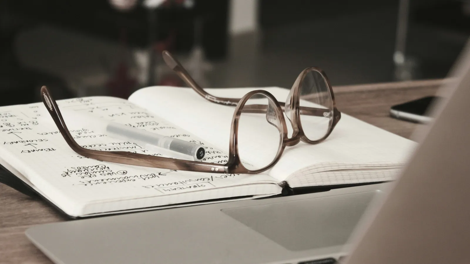 Brown eyeglasses resting on an open notebook with handwritten notes beside a laptop on a wooden desk.