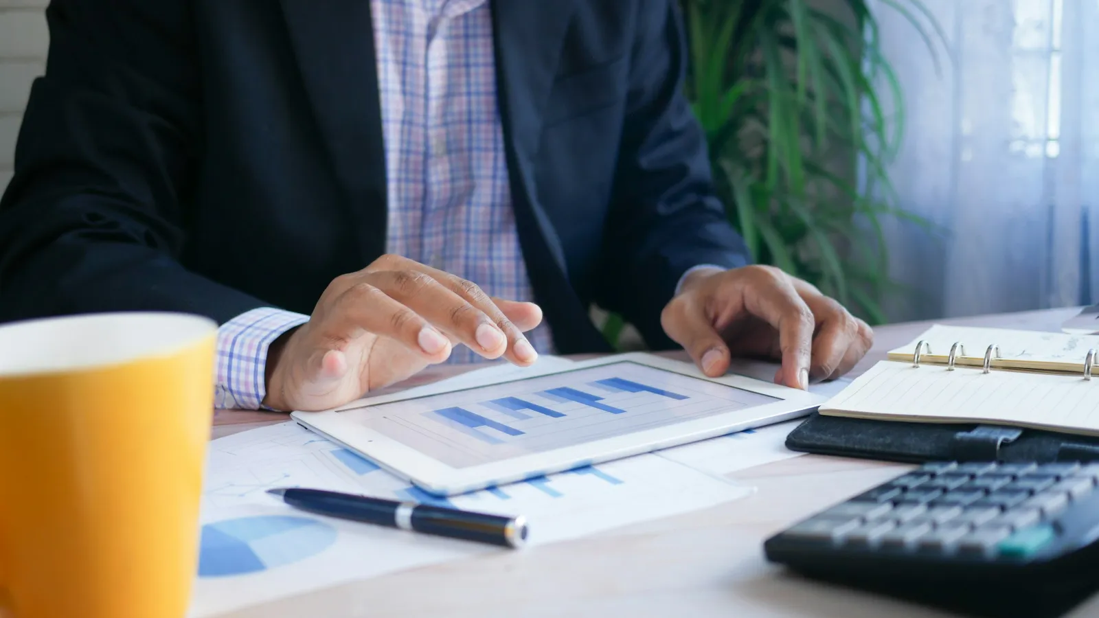 Businessman analyzing bar charts on tablet with calculator, notebook, pen, and coffee cup on desk in bright office.