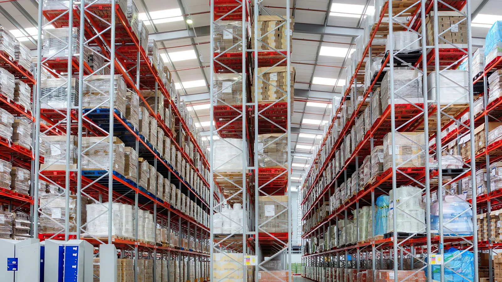 Interior view of a large warehouse with tall shelves stacked with boxes and pallets under bright lighting.