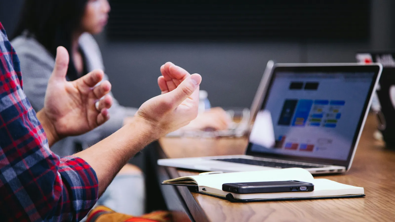 Person gesturing with hands during a meeting with laptop, notebook, and smartphone on the table.