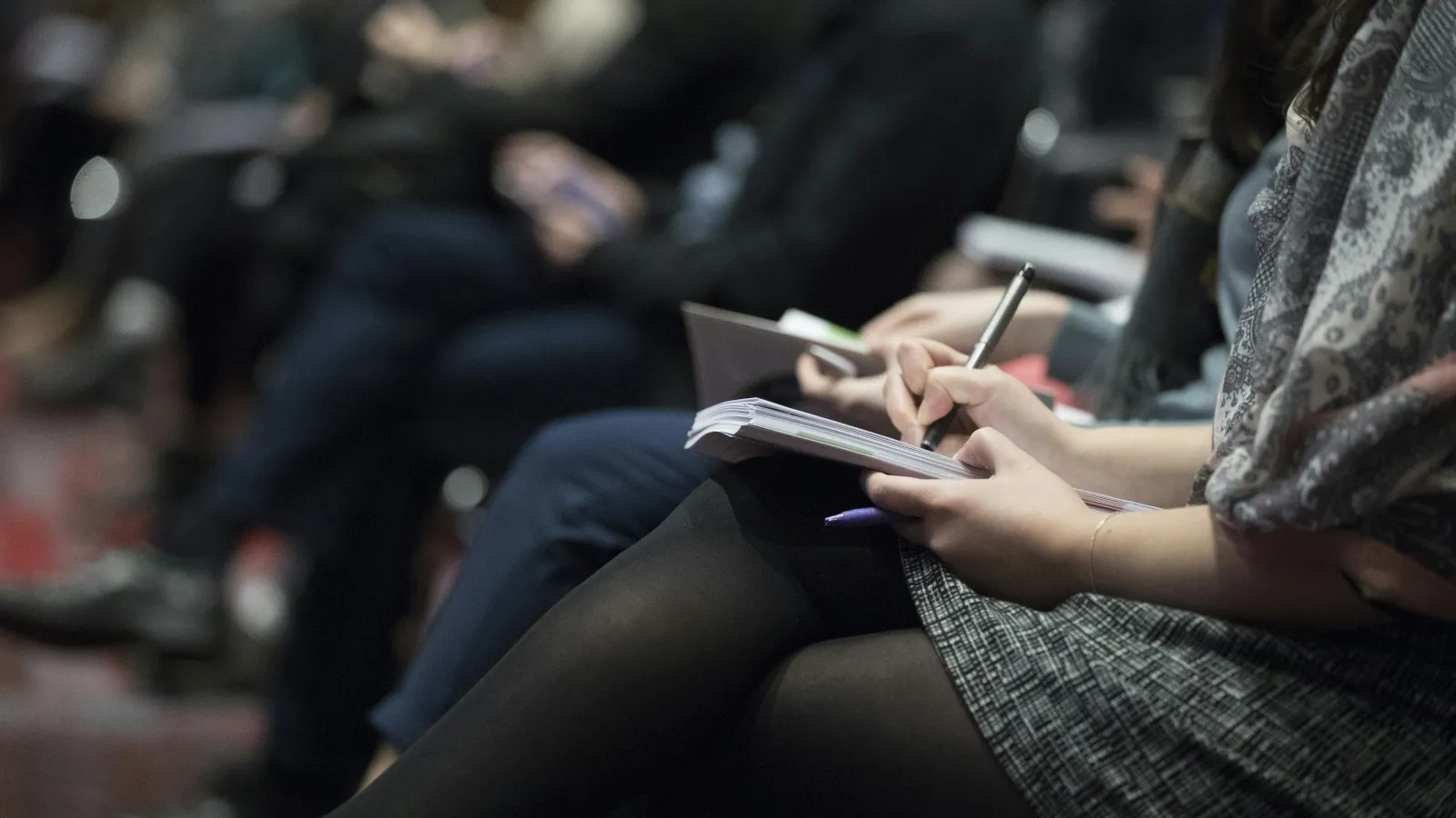 Audience members sitting and taking notes during a seminar or conference in a dimly lit room.