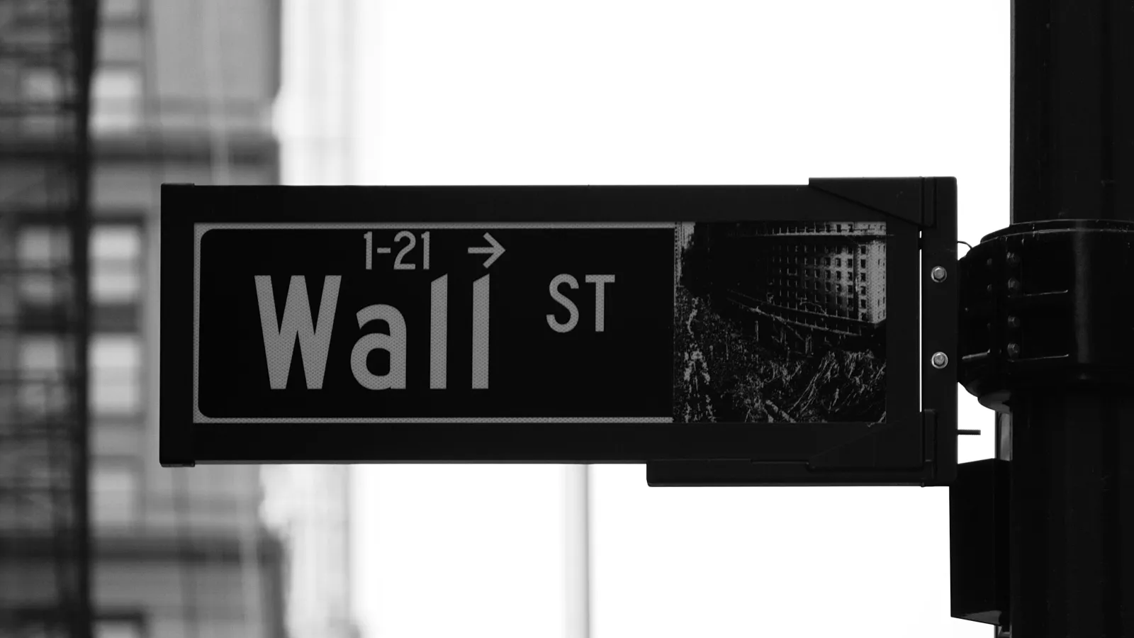 Black and white close-up of Wall Street sign mounted on a pole with blurred city buildings in background.