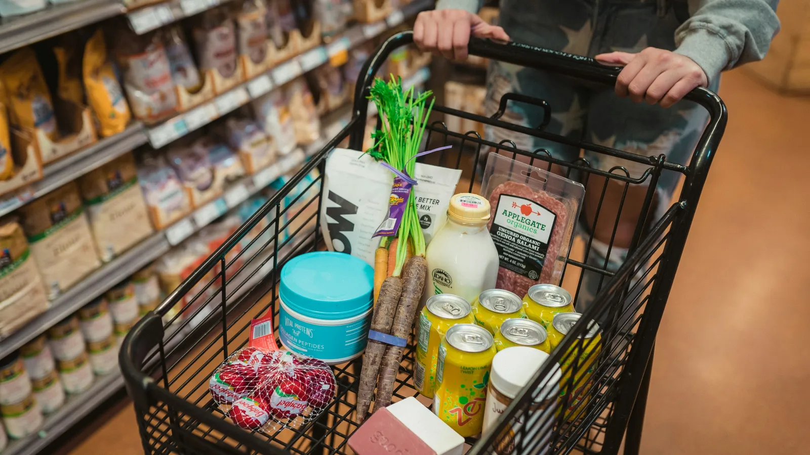 Person pushing shopping cart filled with groceries including drinks, carrots, milk, and packaged goods in a store aisle