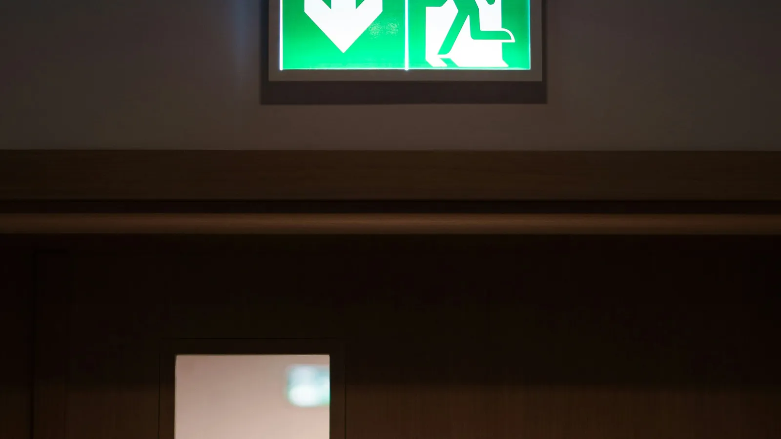 Green emergency exit sign with downward arrow and running figure above a wooden door in a dim corridor.