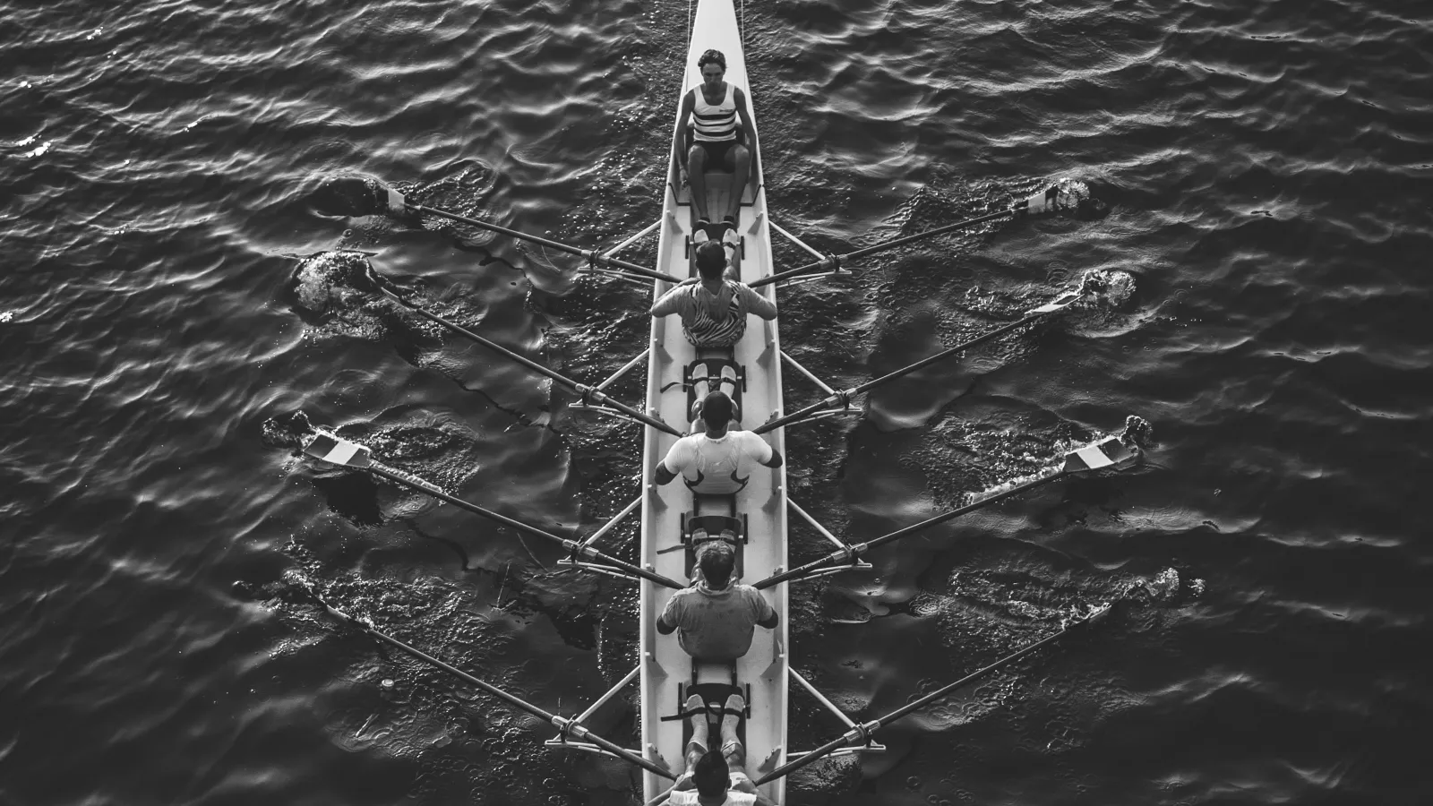 Black and white aerial photo of five rowers in a racing shell rowing on rippled water surface in sync.