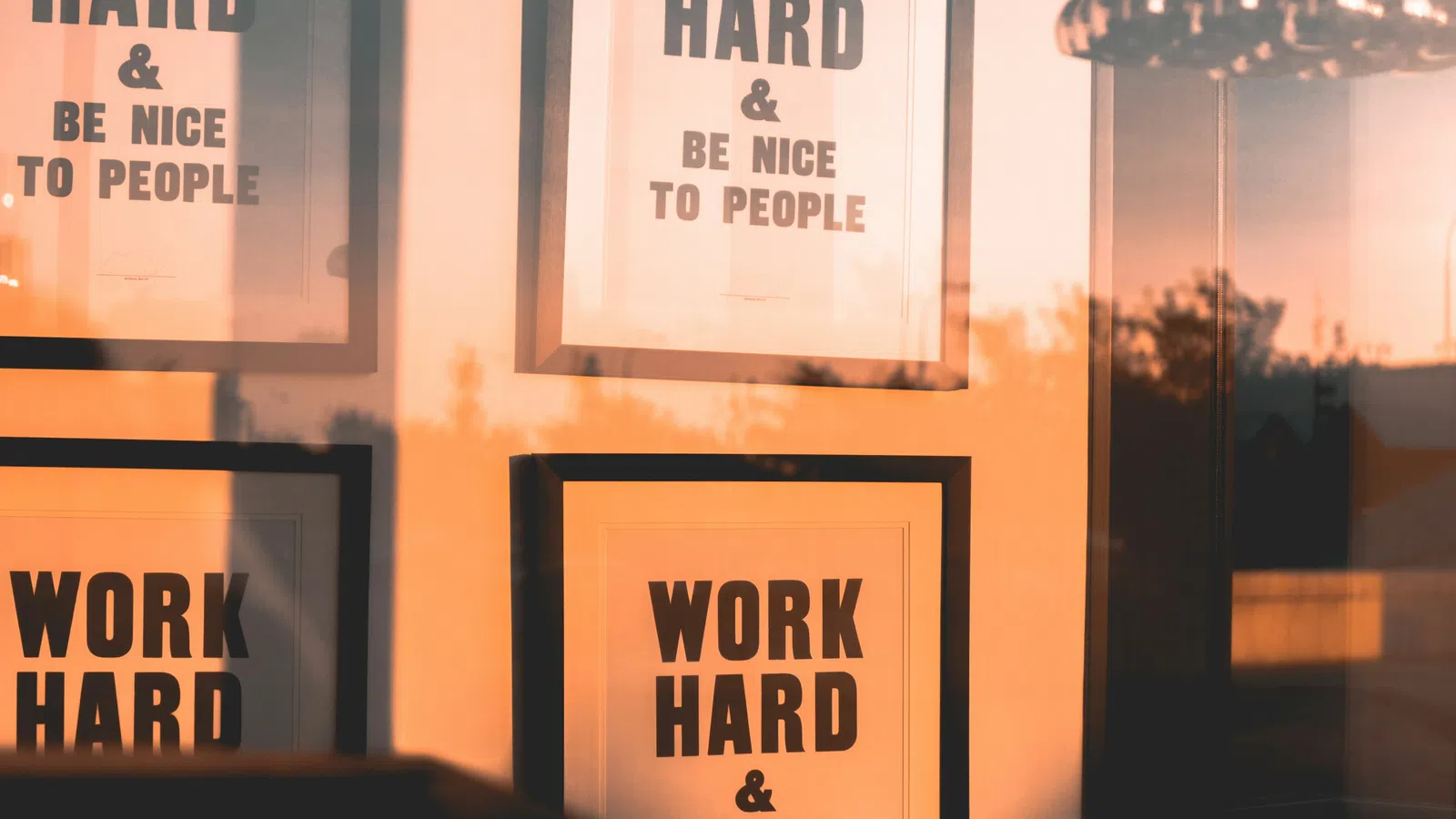 Framed motivational posters reading 'Work Hard & Be Nice to People' reflected in glass during sunset.