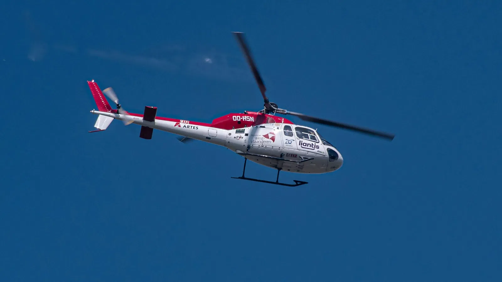 White and red helicopter flying in clear blue sky with visible rotors and landing skis.