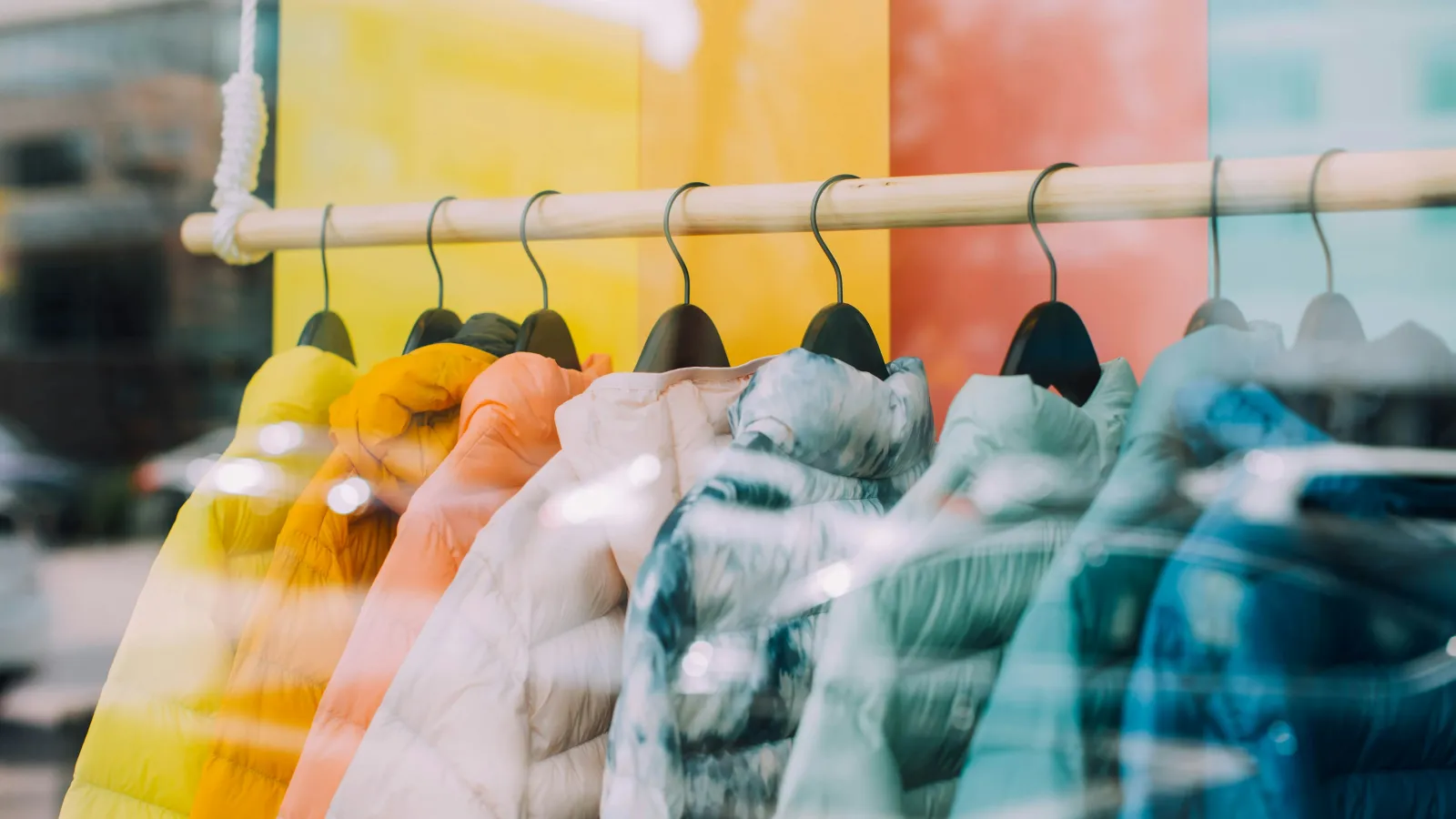 Colorful puffer jackets hanging on a wooden rack behind a shop window with street reflections