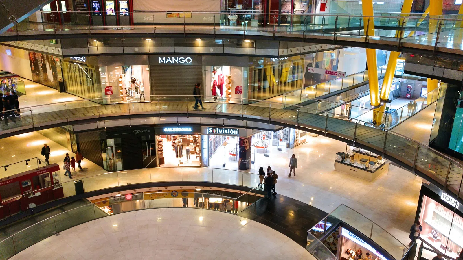 Interior view of a modern multi-level shopping mall with various retail stores and shoppers walking around