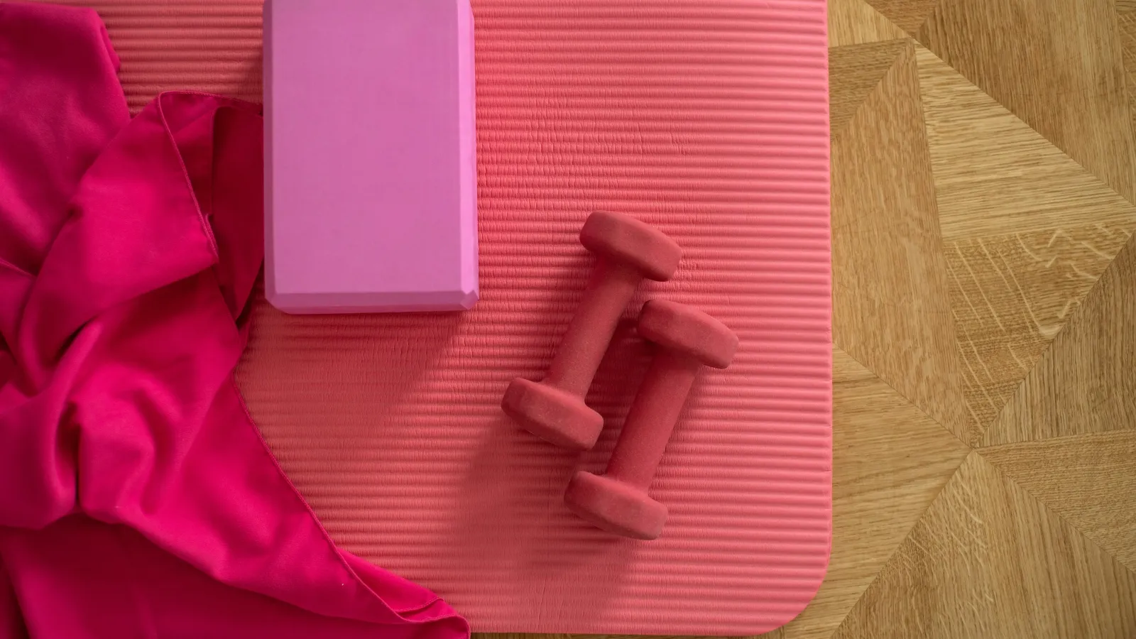 Pink yoga mat with matching dumbbells, a yoga block, and bright pink exercise towel on wooden floor.