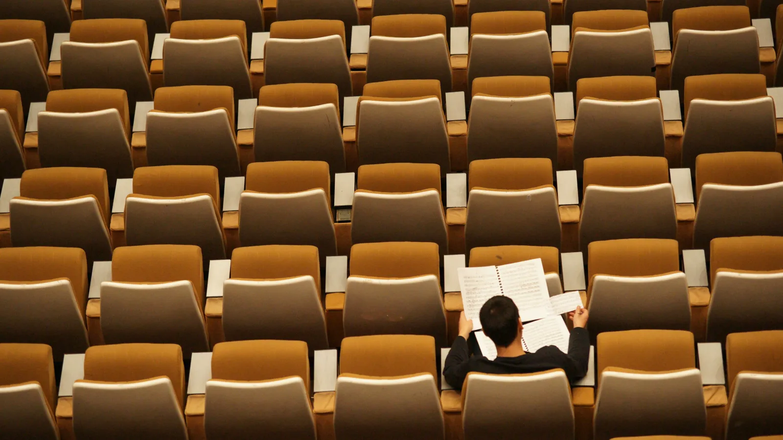 A person sitting alone in a large auditorium reading a music sheet among empty brown seats.