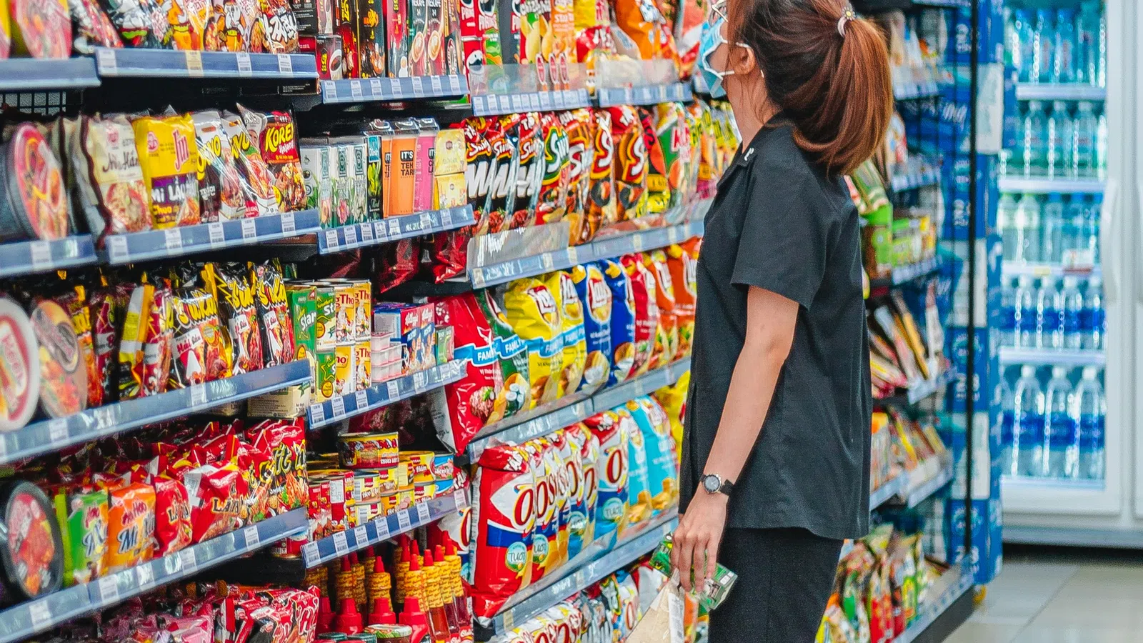 Woman wearing mask shopping for snacks on colorful grocery store shelves in bright aisle.