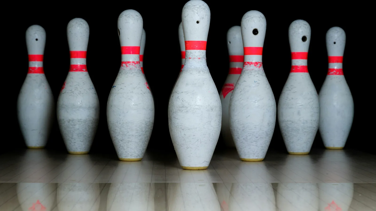 Seven worn white bowling pins with red stripes standing on a polished lane against a dark background