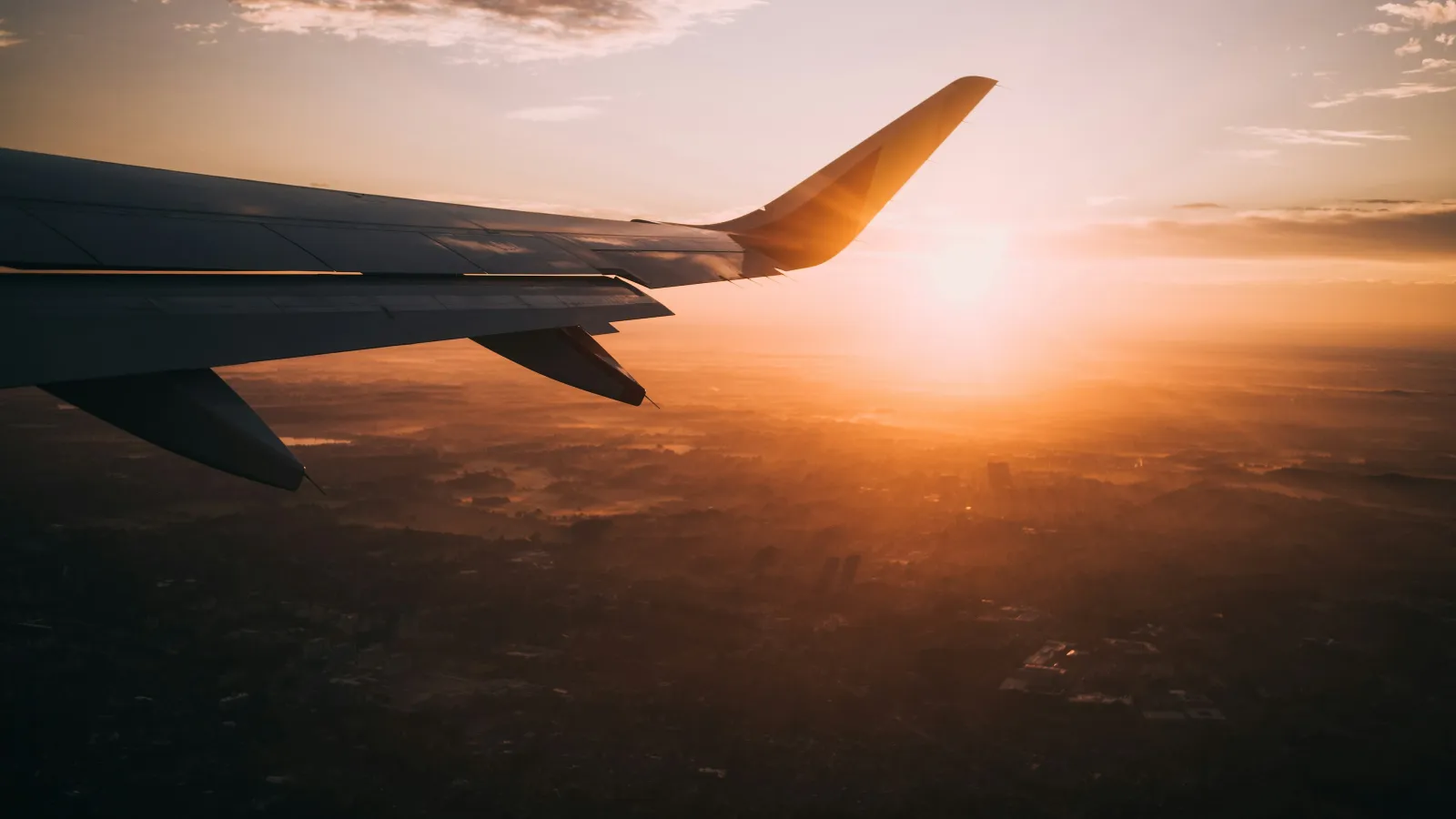Airplane wing above clouds during a golden sunset with a glowing sun on the horizon.