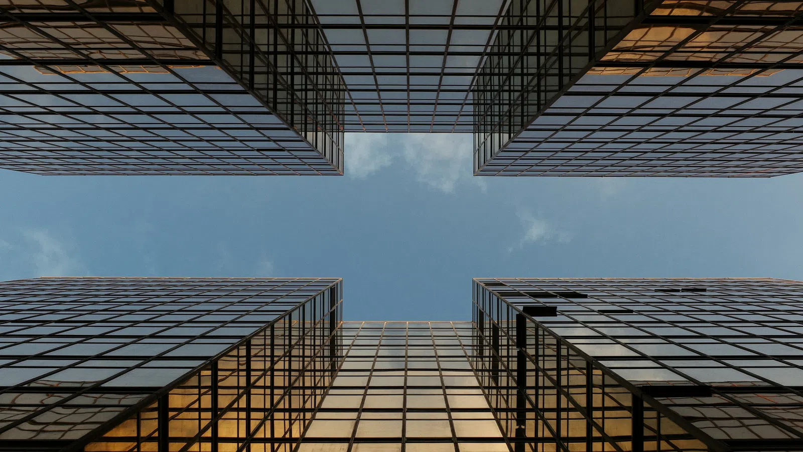 Looking up at modern glass skyscrapers reflecting golden light against a blue sky with clouds.