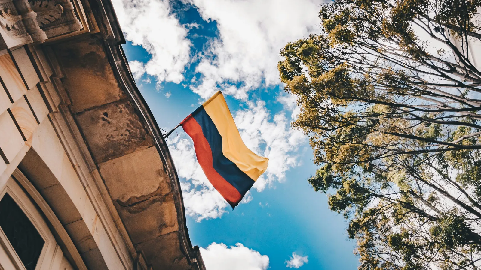 Colombian flag waving against a blue sky with clouds, attached to a historic building with trees nearby.