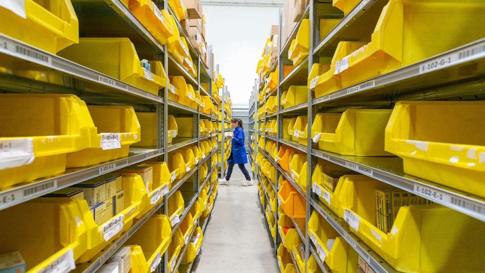 Worker in blue coat walking between rows of shelves filled with yellow storage bins in a warehouse aisle