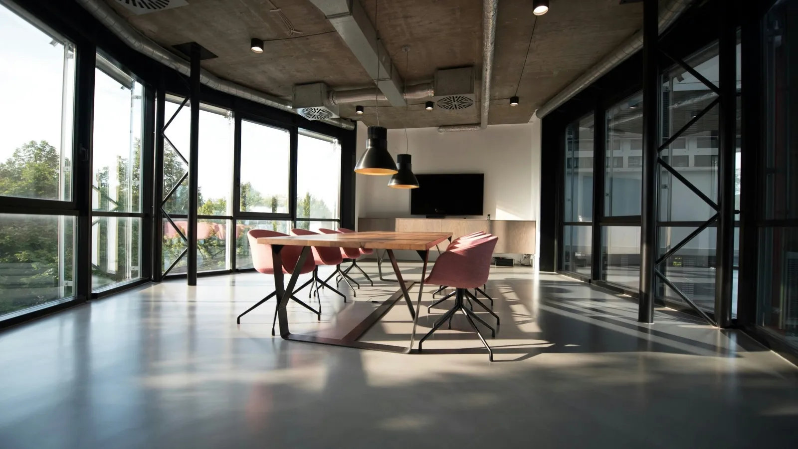 Modern conference room with large windows, natural light, wooden table, pink chairs, and industrial ceiling lights.