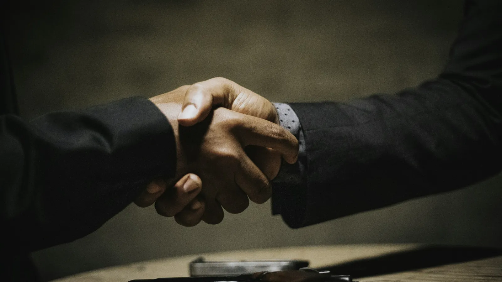 Two people in business attire shaking hands over a table with a handgun and phone visible.