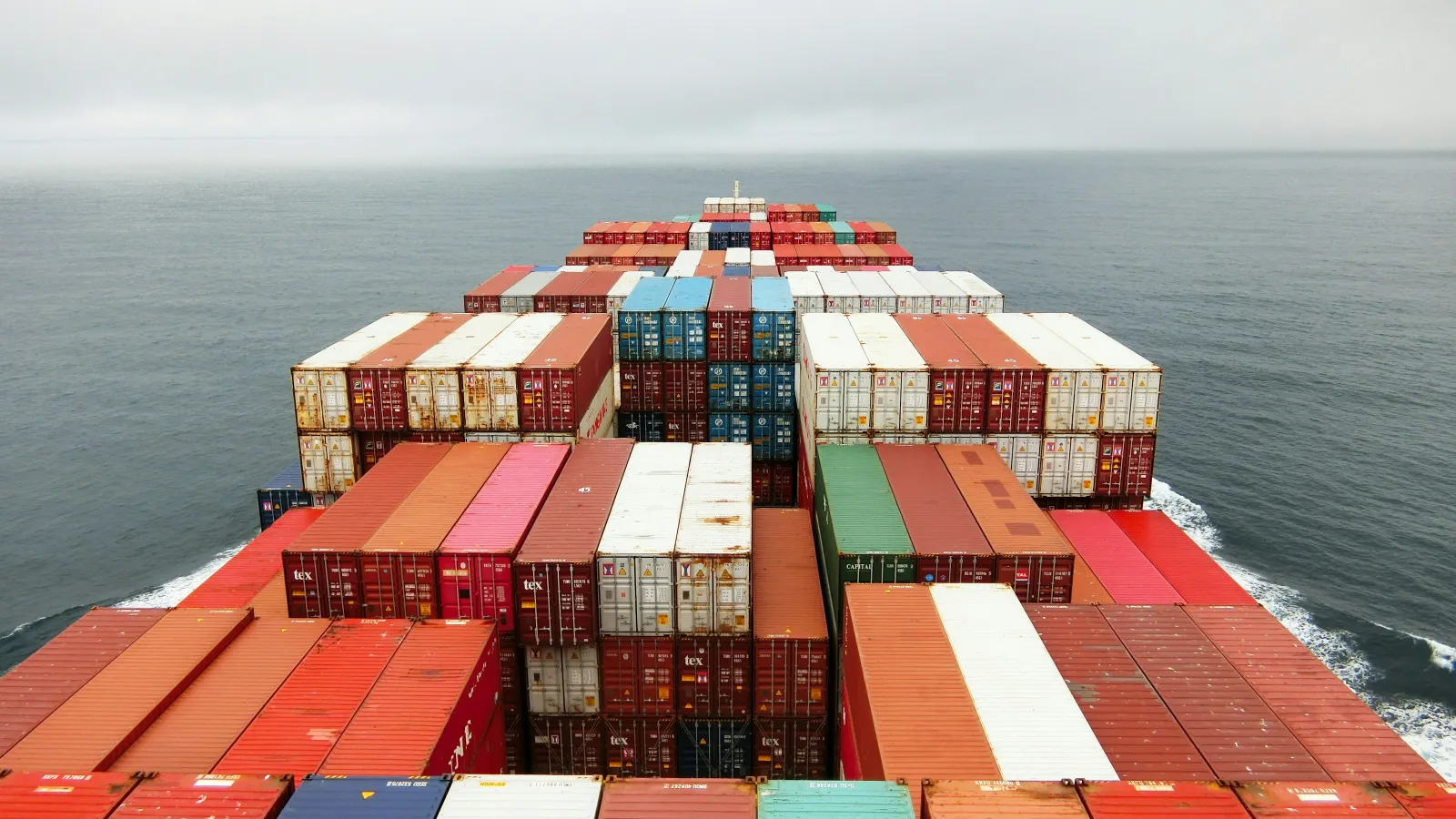 Rear view of cargo ship loaded with colorful shipping containers sailing on calm ocean under cloudy sky.