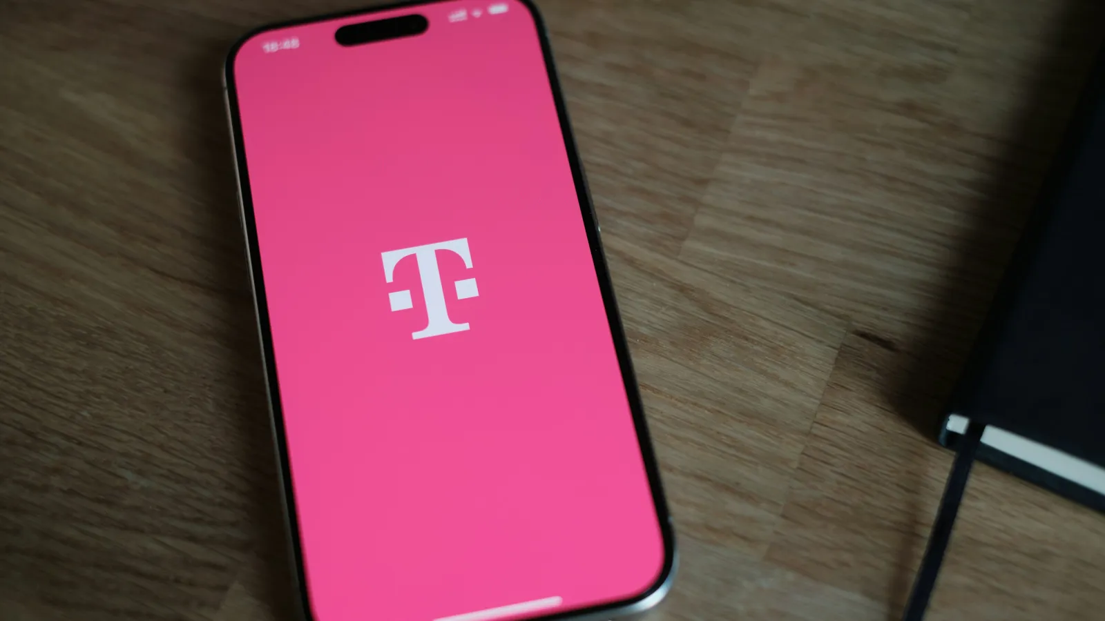 Smartphone displaying the T-Mobile logo on a pink screen on a wooden surface next to a black notebook.