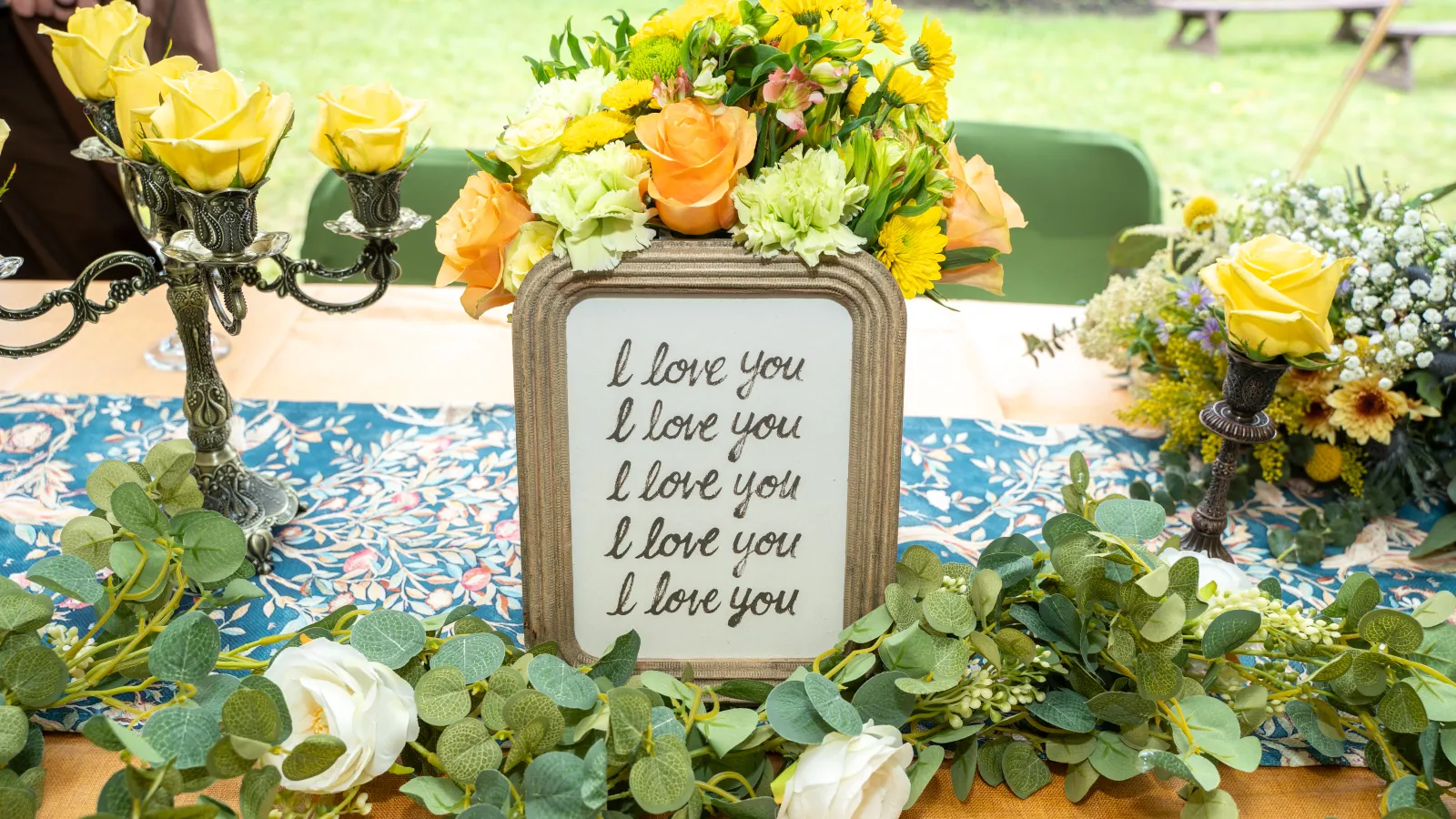 Decorative table with yellow roses, greenery, and a framed sign repeating I love you five times in cursive.