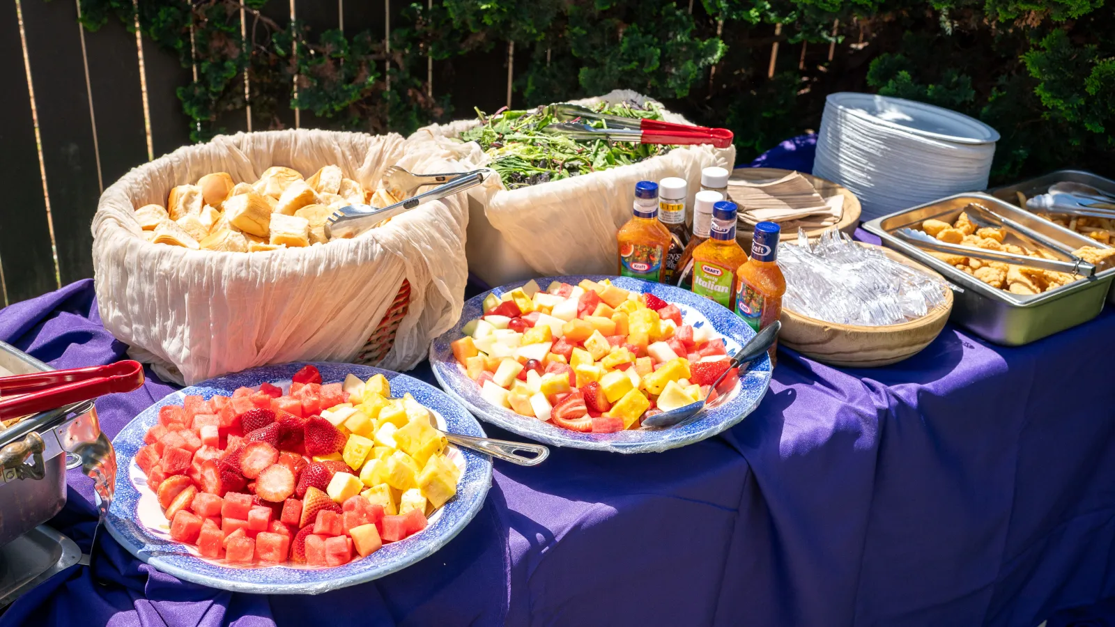 Buffet table with fresh fruit platters, bread rolls, salad, dressings, and plated dishes on a purple tablecloth outdoors.