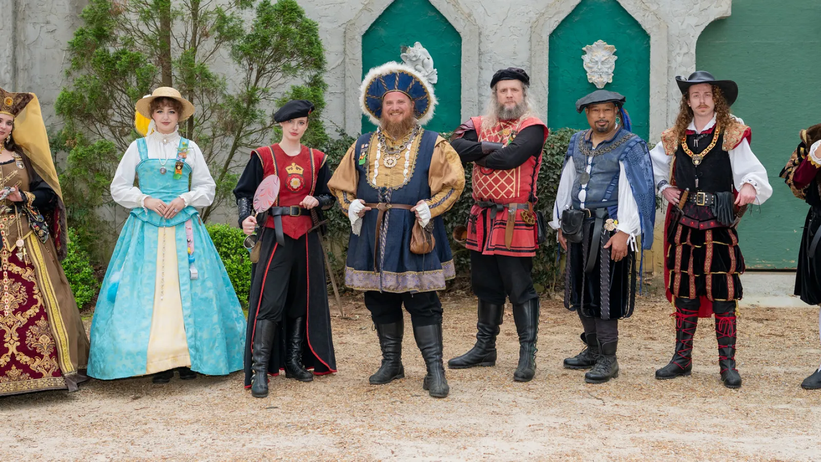 Group of eight people dressed in colorful Renaissance-style costumes posing outdoors against a green and white wall.