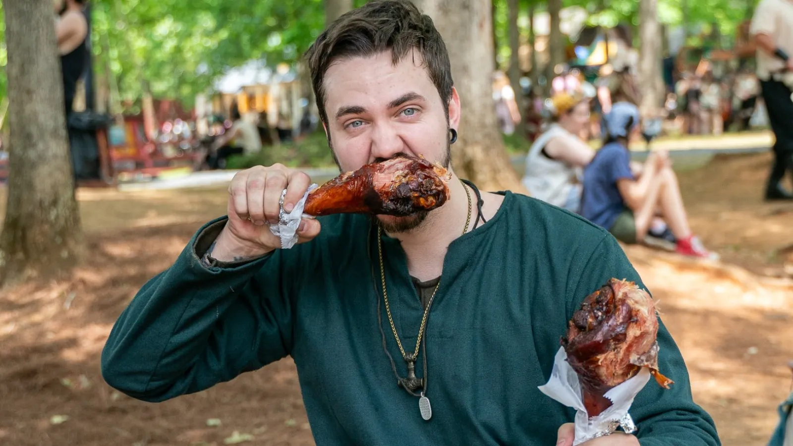Man in medieval costume sitting on bench eating two large turkey legs at outdoor Renaissance fair