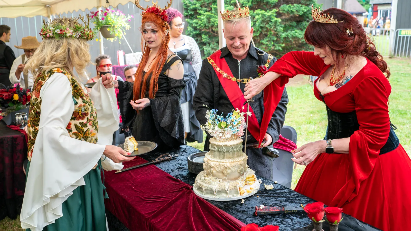 People dressed as medieval royalty cutting and serving a decorated cake at an outdoor event under a tent.