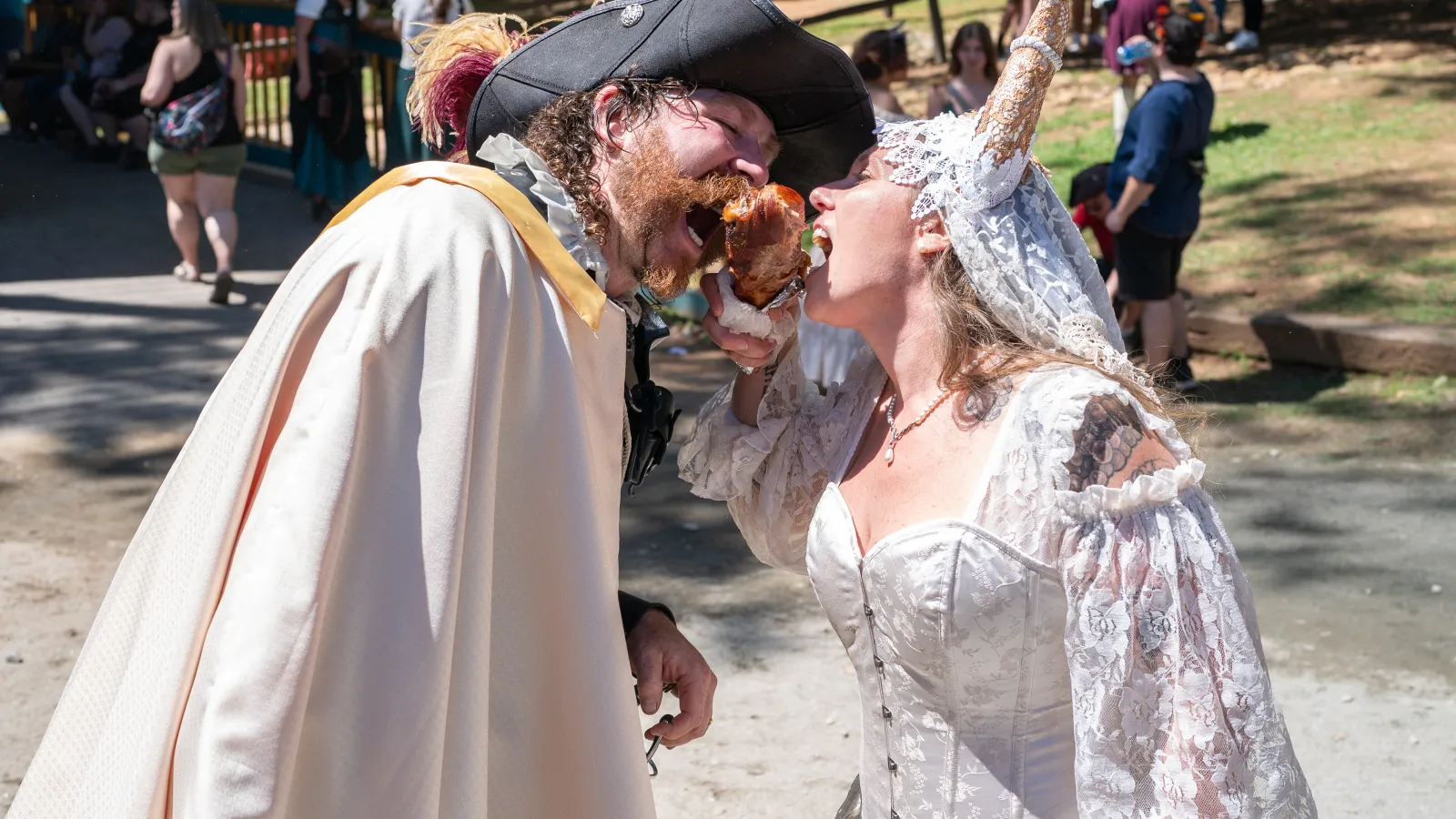 Couple in Renaissance costumes sharing a turkey leg at an outdoor festival on a sunny day