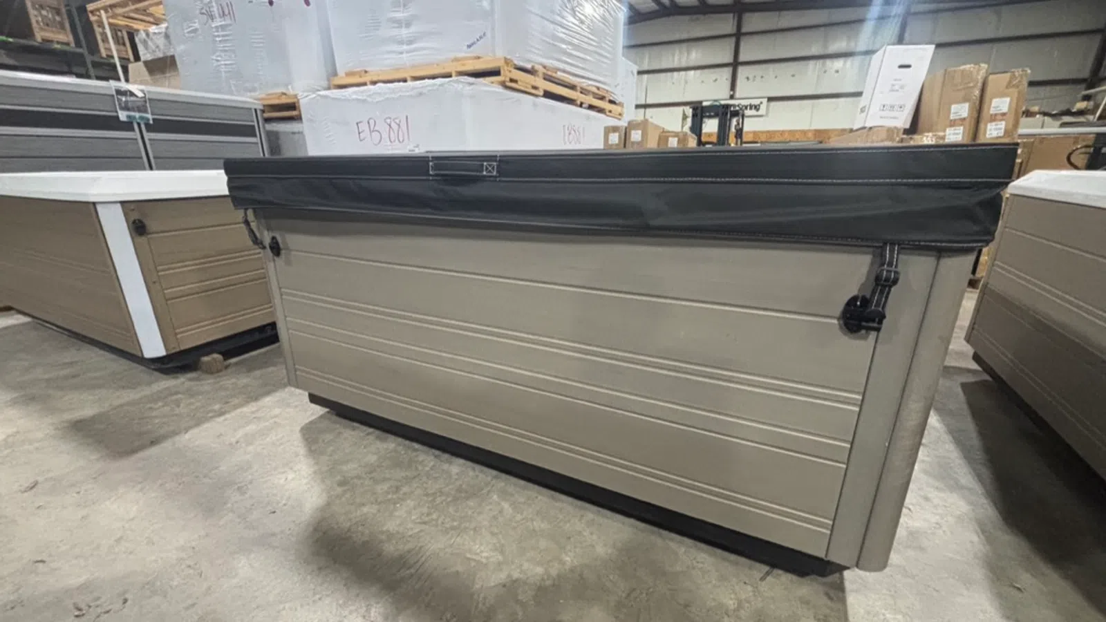 Large beige hot tub with black cover stored inside a warehouse among packaged pallets and boxes.